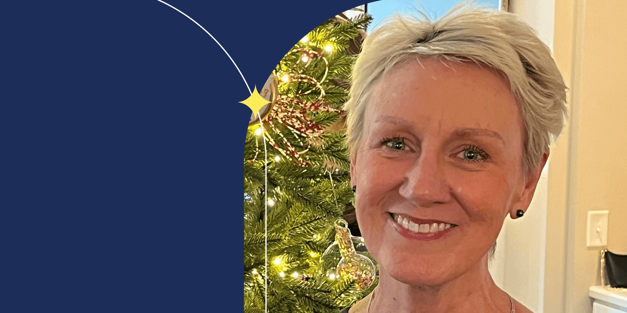 A person facing the camera. They have short hair and blue eyes. They wear earrings and pink lipstick. A Christmas tree can be seen in the background.