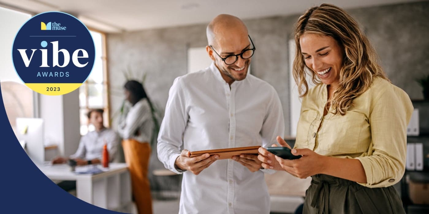 Two coworkers smiling while looking at a phone and tablet in an office.